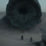 A massive worm is rising its head out of a sand dune while two indistinguishable human figures look in awe.