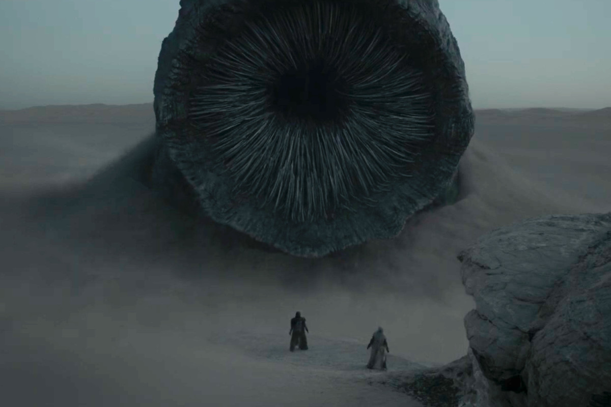 A massive worm is rising its head out of a sand dune while two indistinguishable human figures look in awe.
